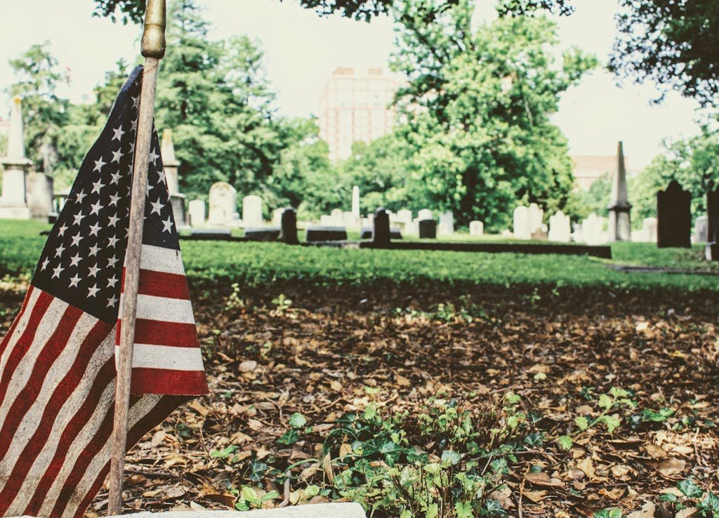 American flag next to tombstones in a peaceful cemetery setting with trees and sunlight.
