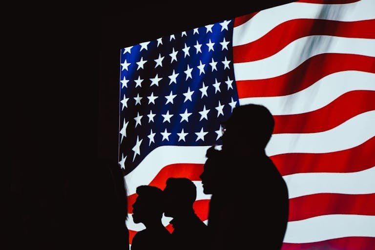 Silhouetted figures in front of a bright, wavy American flag create a patriotic scene.